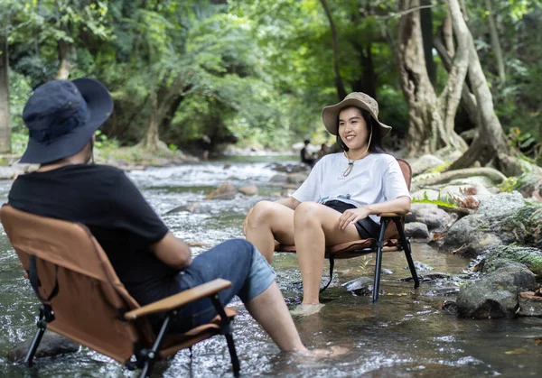 Selective focus of happy woman and man relaxing and enjoying by autumn stream in the forest and sitting on camping chairs while talking together.