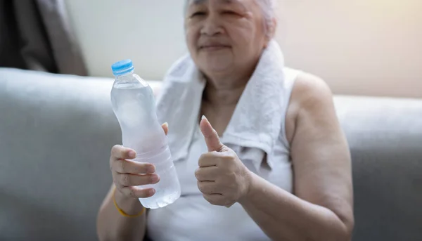 Portrait of a older woman show thump up and a bottled water while sitting on sofa at home. Selective at bottle