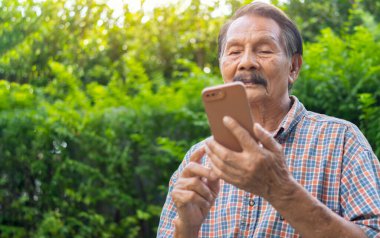 Akıllı telefon kullanıp bahçede dinlenirken kameraya bakan kıdemli Asyalı mutlu bir adam. Modern kablosuz teknoloji eski nesil insan kavramının kolay kullanımı