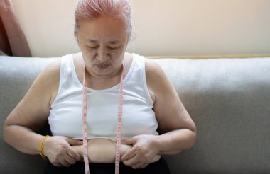 Yaşlı Asyalı şişman kadın, tombul, kilolu, oturma odasındaki koltukta belini ölçen mutsuz kadın diyet yaşam tarzı aşırı kilolu problem konsepti..