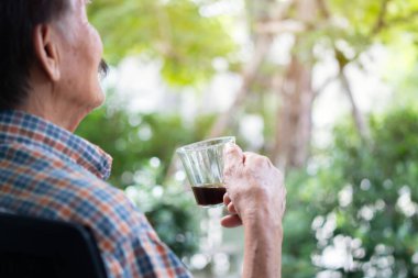Yaşlı Asyalı bir adamın sabah parkın yanındaki sandalyede otururken kahve içmesi. Arka plan, seçici odak.