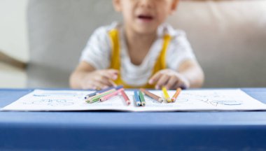 Küçük üzgün çocuk masada oturuyor ve renkli kalemlerle ağlıyor, kapalı bir yerde. İlkokul ve eğitim. Konuya seçici odaklanma.