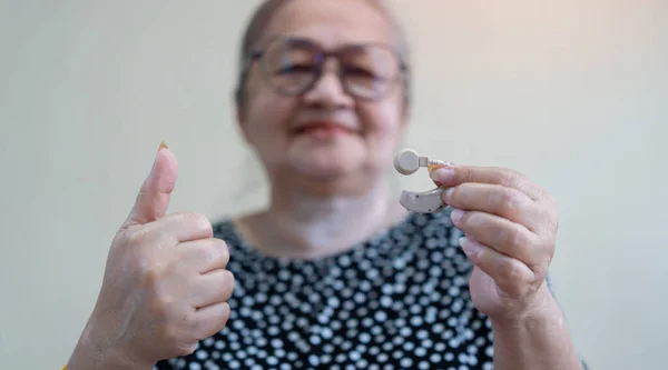 Happy elderly Asian woman holding hearing aid and show the thumbs up on light biege background.  The problem of hearing in old age. Close-up, selective focus.