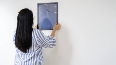 Asyalı kadının çerçevede mavi poster tutuşu ve oturma odasındaki beyaz arka plandaki duvarı dekore edişi, iç mekan kavramı.