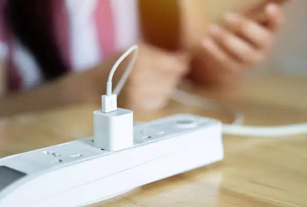 Kadının eli akıllı telefonu tutuyor ve masaya beyaz eklenti prizinden şarj oluyor. Konuya seçici odaklanma.