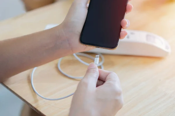 Woman hands plugging a charger in a smart phone with white extension plug on wooden table. Selective focus on hand.