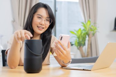 Mutlu Asyalı kadın cep telefonu kullanırken evde ses asistanıyla konuşuyor. Akıllı yapay zeka hoparlörü. Kıza seçici odaklanma.