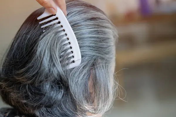 Middle age grey-haired woman on back view combing hair standing at home. Gray hair concept, selective focus.
