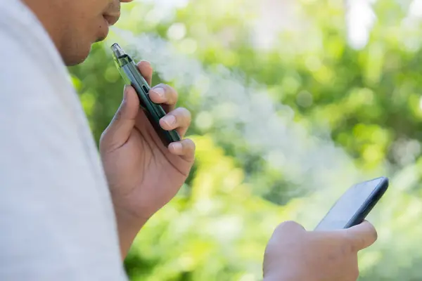Genç adam dışarıda elektronik sigarayla buharı içine çekiyor. Vape aromalı sıvı kimyasallar. Yan görünüm, kapat.