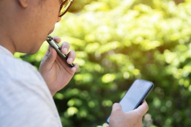 Genç Asyalı adam halka açık alanda buhar çekiyor. Parkta elektronik sigara içiyor. Nikotin bağımlılığı.