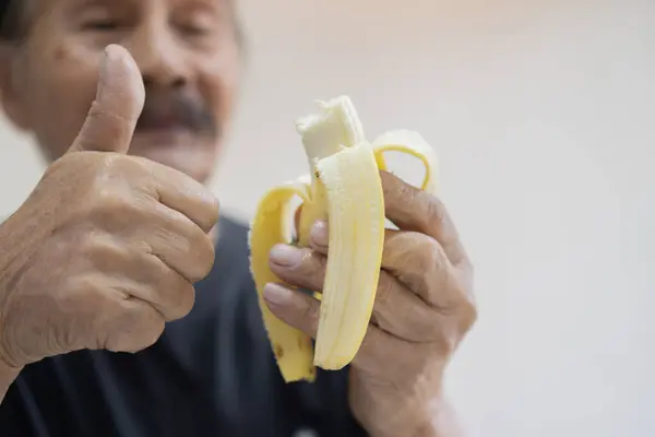 Defocused happy senior man wwith thumb up and isolated on a white background holds a peeled banana in his hand ready to be eaten