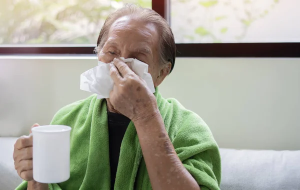 Shot of old age Asian man feeling sick with cold and fever at home, ill with flu disease sitting on the sofa and drink warm water.