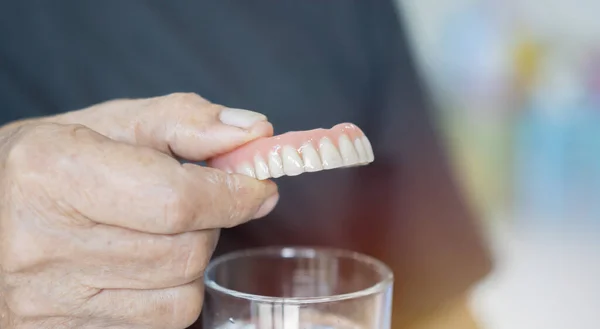 Close up of false teeth in old man hand over the glass. Old age, removable denture concept. Selective focus, copy space.