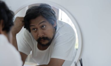 Sakallı yakışıklı, orta yaşlı Asyalı adam aynaya bakıyor. Saç dökülmesinden endişelenen orta yaşlı bir adam. Erkeklerin kişisel bakımı. Sabah rutini