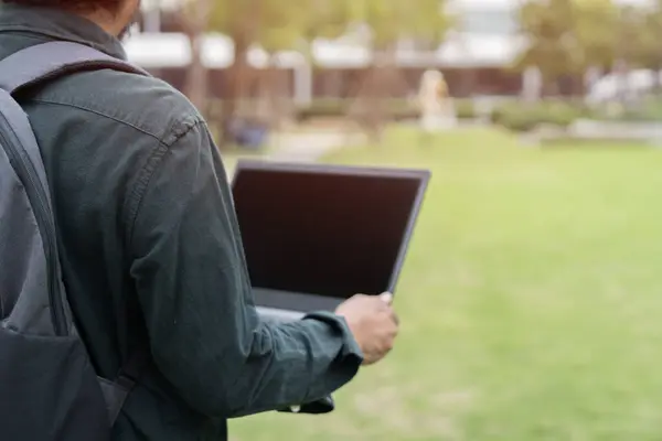 Back view of man standing on grass at park working on laptop. Male wearing backpack and holding laptop with bright sunlight.