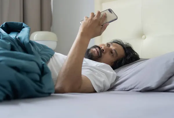 The serious Asian man lay on the bed and phone at morning in bed. Side view, selective focus.