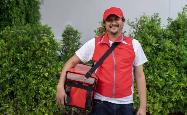 Smiling delivery employee man in red cap t-shirt uniform thermal food bag backpack work courier service standing over nature background
