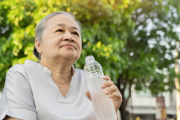Old Asian woman drinks water after walking exercise in the park. The concept of a healthy lifestyle. Copy space