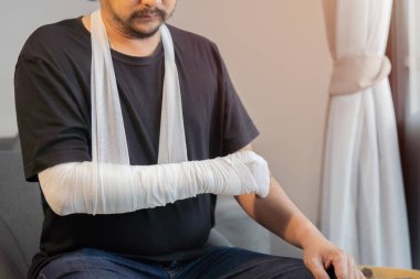 Sakallı Asyalı adam, yaralı dirseğiyle beyaz bandajlı gazlı bezle evde oturuyor. Dirsek yarası.