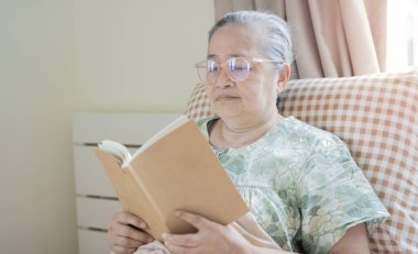 Tazelenmiş 60 'lı yaşlarda bir kadın sabah uyanıyor, kitap okuyor, yatak odasında evde oturuyor.