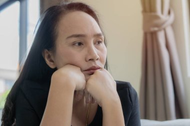 Oturma odasındaki kanepede oturan depresif Asyalı kadın ve evdeki pencereden bakan. Seçici odak.