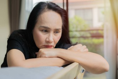 Yetişkin bir Asyalı kadın evdeki kanepede oturup pencereye bakıyor. Hayal kırıklığına uğramış kadın, özel hayatında mutsuz bir sorun hissediyor. Boşluğu kopyala.
