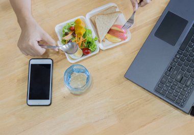 Laptop ve cep telefonuyla beyaz tahta masada lezzetli sağlıklı salata yiyen bir kadın. Üst görünüm