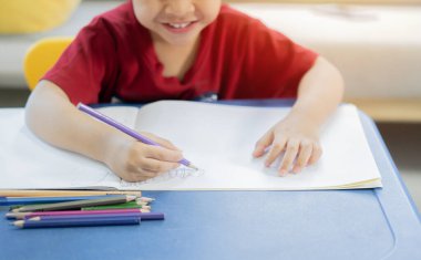 Evdeki sanat kitabının üstünde kalem ve renkle resim yapan sevimli, mutlu, küçük Asyalı bir çocuğun görüntüsü. Yaratıcı genç bir sanatçı iş başında. Çocuklar resim yapar. Çocuklar çizer.