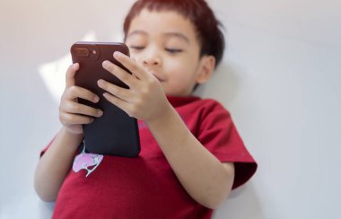 Kırmızı tişörtlü küçük bir çocuk yalan söylüyor ve cep telefonu akıllı telefonunda uygulama kullanıyor. Seçici odak.