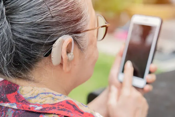 İşitme cihazları, sağırlık tedavisi, işitme biliminde yenilikçi teknolojiler. İşitme cihazı olan olgun bir kadın dışarıda akıllı telefon kullanıyor..