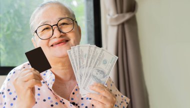 Çevrimiçi alışveriş, pazar yeri başvuru kullanıcısı. Yaşlı gülümseyen Asyalı kadın elinde Monny ve bankamatik kartıyla evdeki kanepede oturup rahatça e-alışveriş yapıyor..