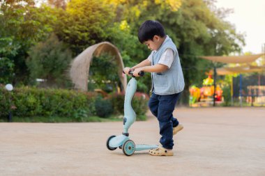 Çocuk güneşli bir yaz gününde parkta scooter sürmeyi öğreniyor. Anaokulu çocuğu paten kayıyor. Çocuklar dışarıda scooterlarla oynarlar..