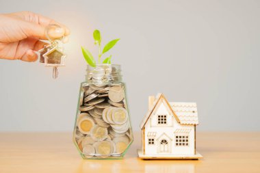 Elinde para kavanozu bozuk paralar ve ahşap masada ev modeli olan ev anahtarı. Yatırım ve emlak kavramı, ev konsepti almak için para biriktirmek.