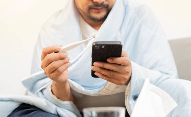 Grip hastası bir adam termometreyi tutuyor ve evdeki telefonuyla grip hakkında tıbbi bilgilerle ısıyı kontrol ediyor. Seçici odak.