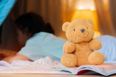 Kahverengi bir oyuncak ayı açık bir kitabın üzerine oturur ve arkasında masaların altında oynayan küçük şirin bir kızın bulanık görüntüsü vardır. Çocuklar Oyun Evi Konsepti.