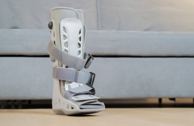 Bir ortopedik gri plastik çizme bilekliği yaralanması. Yakınlarda çizmeleri koruyor. Arka planda gri bir kanepe var. Kırık ayak koruması..