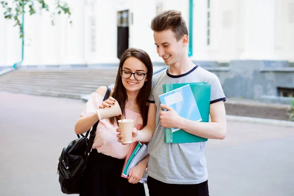 Two friends students near university with coffee. Attractive girl share some coffee with her handsome classmate, ready to help each other in difficult situation. Concept of friendship.