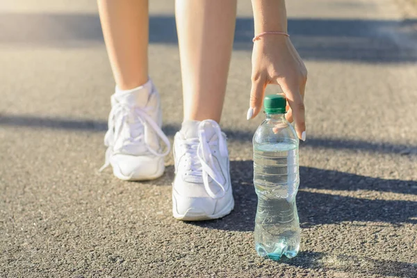 Athletic girl in white sneakers stopped on the road in order to take a pause and drink some water out of plastic bottle during jogging workout in the evening.