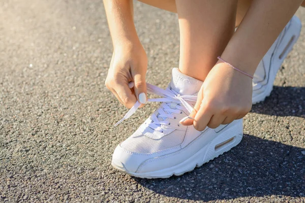 stock image Sportsmen girl in white sneakers stopped on the road and typing shoelaces while jogging workout in the evening. Closeup of female feet