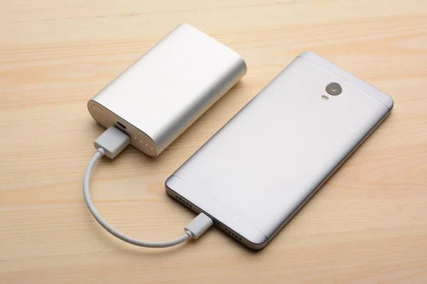 現代の銀製スマートフォンは パワーバンクと充電しながら その画面をライト木製のテーブルの上に置く — ストック写真