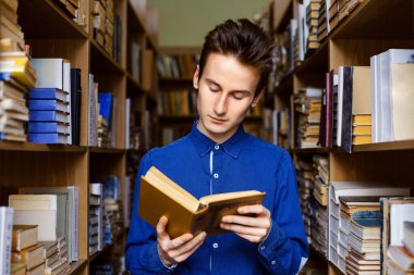 Kütüphanede kitap okuyan bir erkek öğrenci, farklı eski kitapların raflarının arasında duruyor.