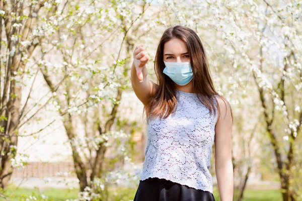 彼女の刺激を示すために 下に親指を示す開花公園に立っているとコロナウイルスの拡散を防ぐために 一定の着用医療用マスクに嫌い — ストック写真