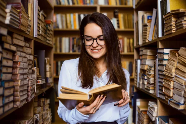 陽気ブルネット女性学生読書本で図書館 異なる古い本の棚の通路の間に立って — ストック写真