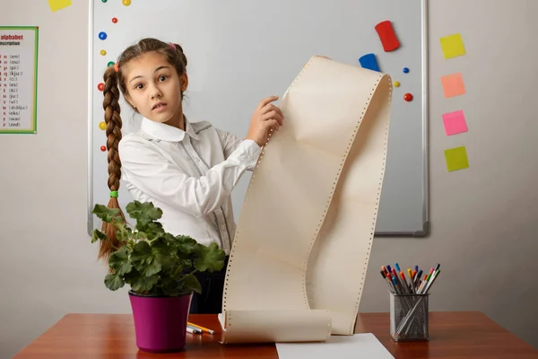 Shot of little girl in school uniform, standing near the desk, looking shocked to the camera, holding a long list of homeworks. Human facial expressions, emotions, education, school.