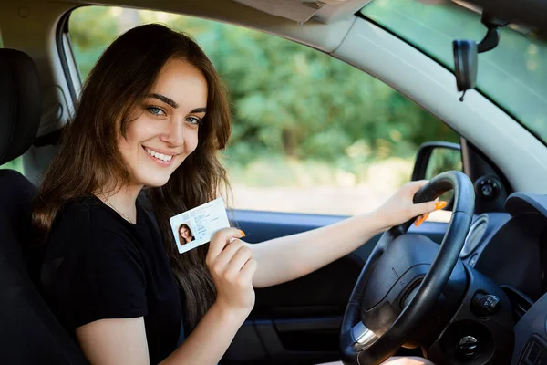 笑容可亲的年轻女子骄傲地展示了她的驾驶执照 坐在新车里 还是个缺乏经验的年轻司机 满面春风 — 图库照片