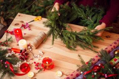 Köknar ağacı dalları ve şenlikli süslemeleri olan açık ahşap masa. Bir kadının elinde Noel çelengi.