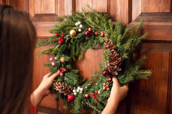 Evinin kapısına Noel çelengi asan genç bir kızın arka görüntüsü. Kış tatili için hazırlık, ev dekorasyonu, sembol.