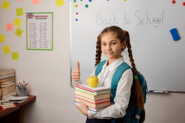 Bir yığın kitabı, elması ve sınıfta duran büyük okul çantasıyla baş parmağını gösteren genç bir kız. Mutlu liseli kız, çalışma zamanı.