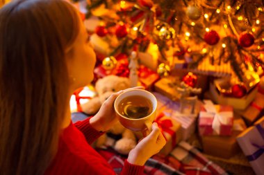 Genç sarışın kız, Noel ağacının güzelliğine hayran. Altında bir sürü hediyeyle evde bir fincan çayla. Noel tatili, şenlikli ev atmosferi