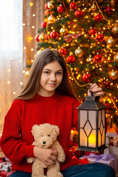 Girl with teddy bear and lantern sitting under Christmas tree. Happy teenage girl at Christmas morning checking presents